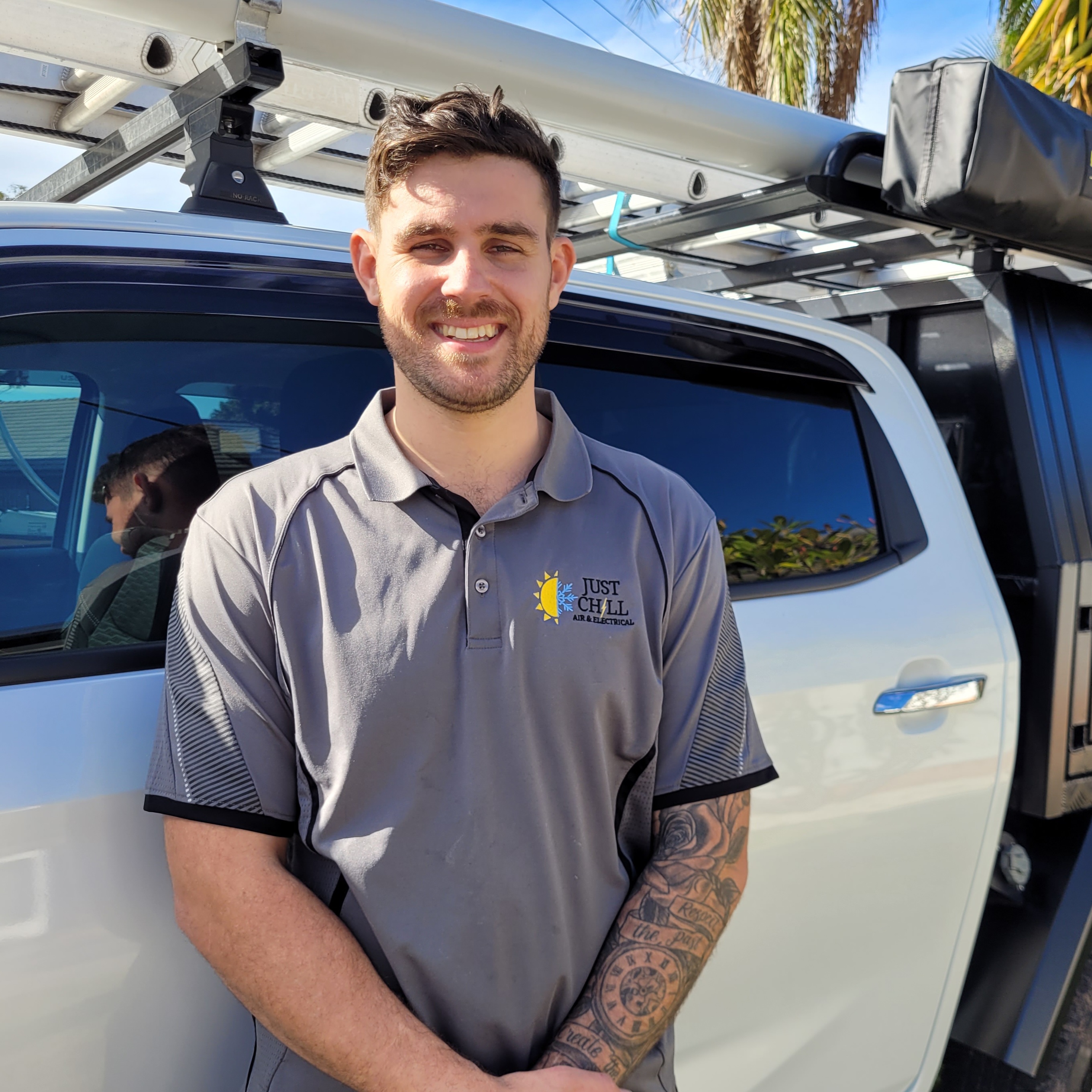 A young man with short brown hair is smiling, standing in front of a vehicle. He is wearing a grey polo shirt with a logo on it. The vehicle is parked in a sunny outdoor setting, surrounded by palm trees.