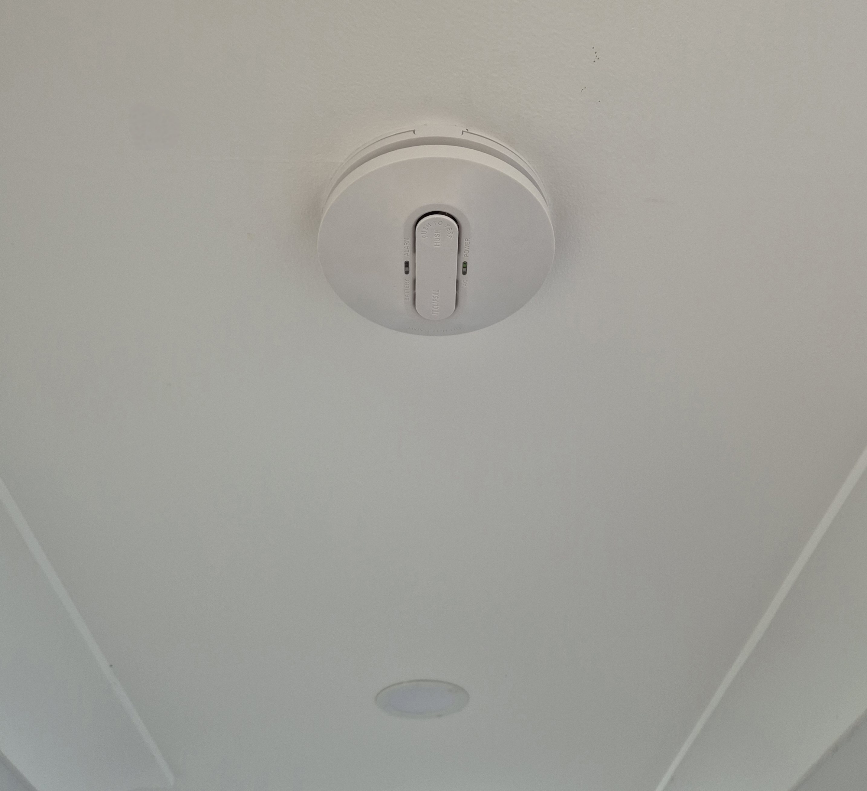 The image shows a ceiling-mounted device in a bright, white ceiling. This device appears to be a smoke detector or a similar safety device, which is important for ensuring safety in environments with air conditioning systems. Proper installation of safety devices is crucial in settings that involve the use of air conditioning to prevent risks associated with fire hazards and to ensure compliance with local regulations. Just Chill Air & Electrical, as an air conditioning contractor, would advocate for the integration and maintenance of such safety devices in their installations and services to enhance overall safety for their clients.