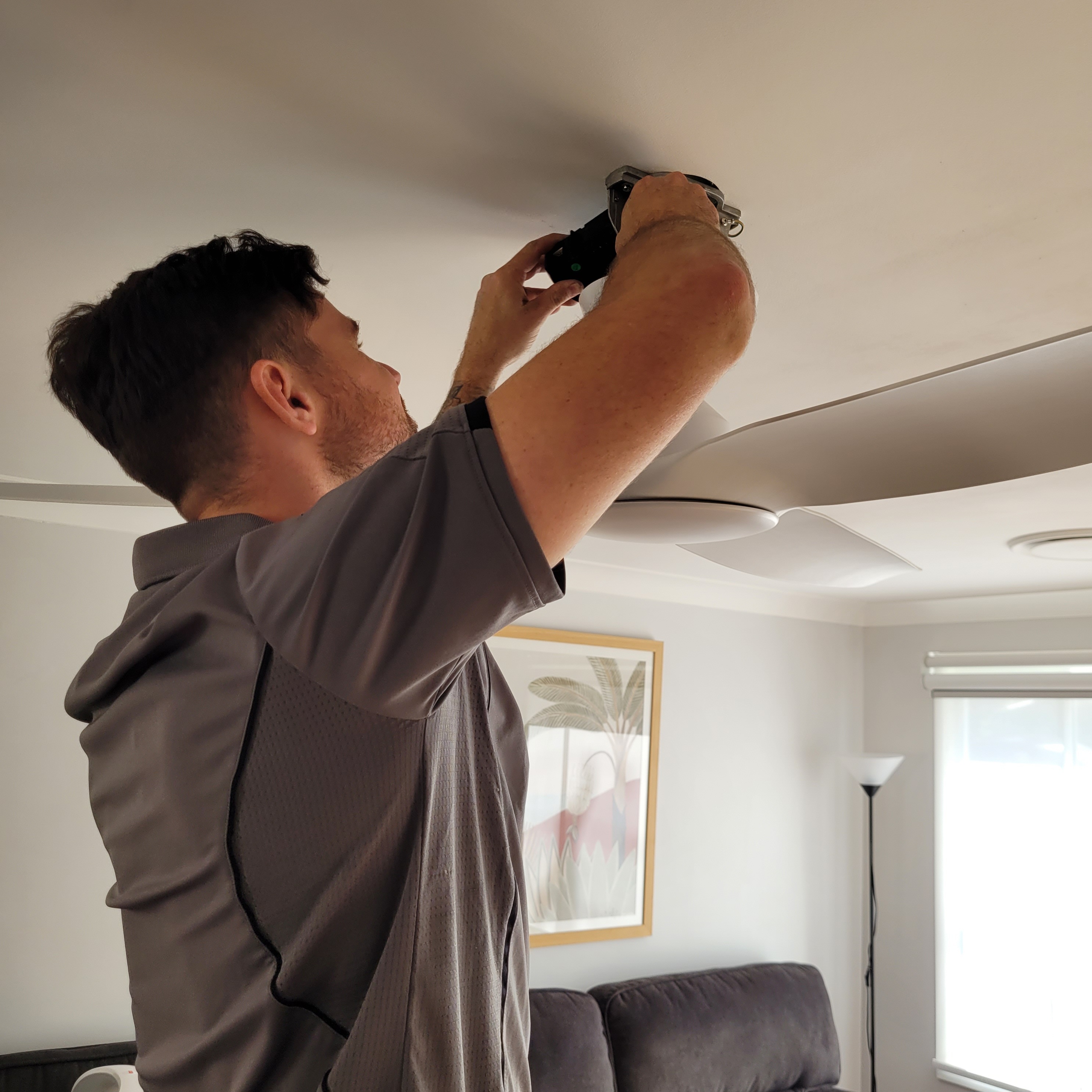 A person is standing on a ladder, working on the ceiling of a room. They are using a power tool to install or repair something, possibly related to air conditioning or ventilation. The room features modern decor with a sofa, a lamp, and a painting visible in the background.