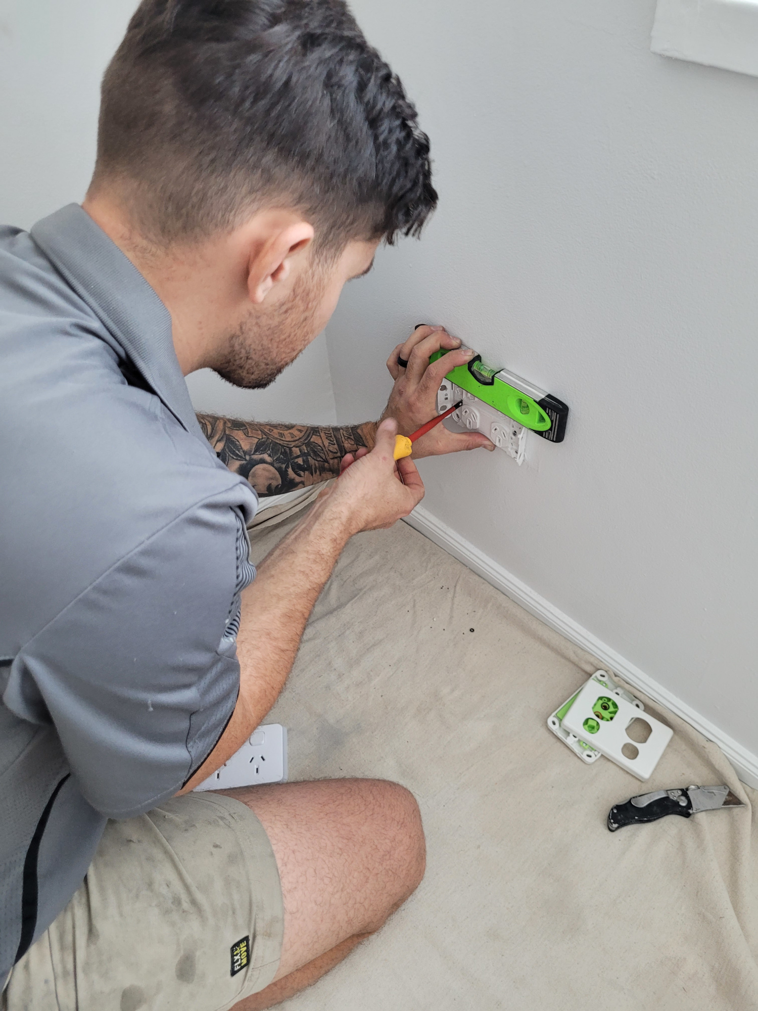 The image shows an air conditioning technician, possibly from Just Chill Air & Electrical, working on installing or repairing an electrical outlet on a wall. The technician is focused and using a level tool, ensuring that the installation is accurate. Their attire is professional, featuring a grey shirt with the sleeves rolled up and shorts appropriate for a work environment. The area around them is covered with a drop cloth to protect the flooring. The electrical outlet being worked on is black, which may indicate a modern design. This work reflects the technician's expertise in electrical and air conditioning systems.